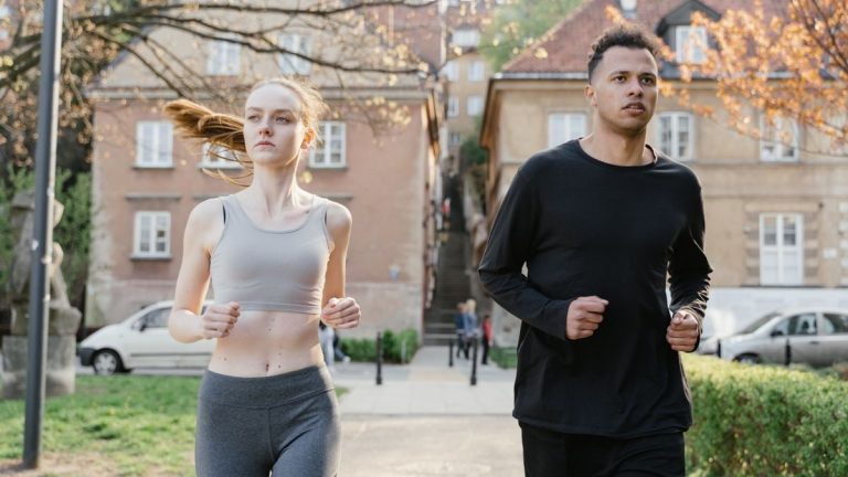 A young woman and man jogging on a city street during the day, wearing sportswear.